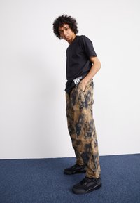 Black V-neck t-shirt, tan and black tie-dye loose-fit pants, and black sneakers. Notable details include a logo patch on the pants.