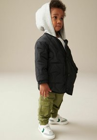Svart vadderad jacka med en grå huvtröja, gröna cargobyxor och vita sneakers med grå och gröna detaljer. Texturerad tyg och synliga fickor.