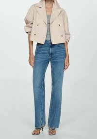 Veste beige croppée à double boutonnage avec de gros boutons dorés, associée à un jean bleu clair droit et des sandales à talons transparents.