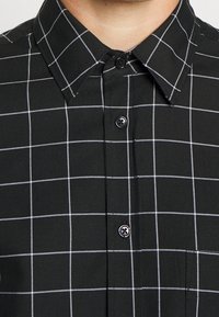 Camicia nera con bottone e motivo a griglia bianca. Presenta un colletto classico e bottoni neri. Il materiale sembra essere tessuto intrecciato.