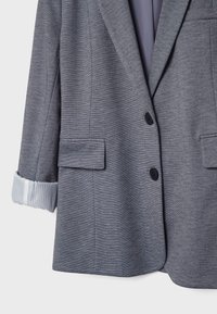 Veste grise en tissu texturé, avec deux boutons noirs et une poche poitrine. Les poignets dévoilent une doublure blanche à rayures bleues.