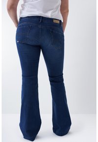 Personne portant un jean évasé bleu foncé et une chemise blanche, vue de dos sur un fond clair uni.