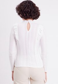 Kvinde med krøllet mørkt hår iført en hvid langærmet bluse i blonde med ruffles og beige bukser, som vender væk på hvid baggrund.