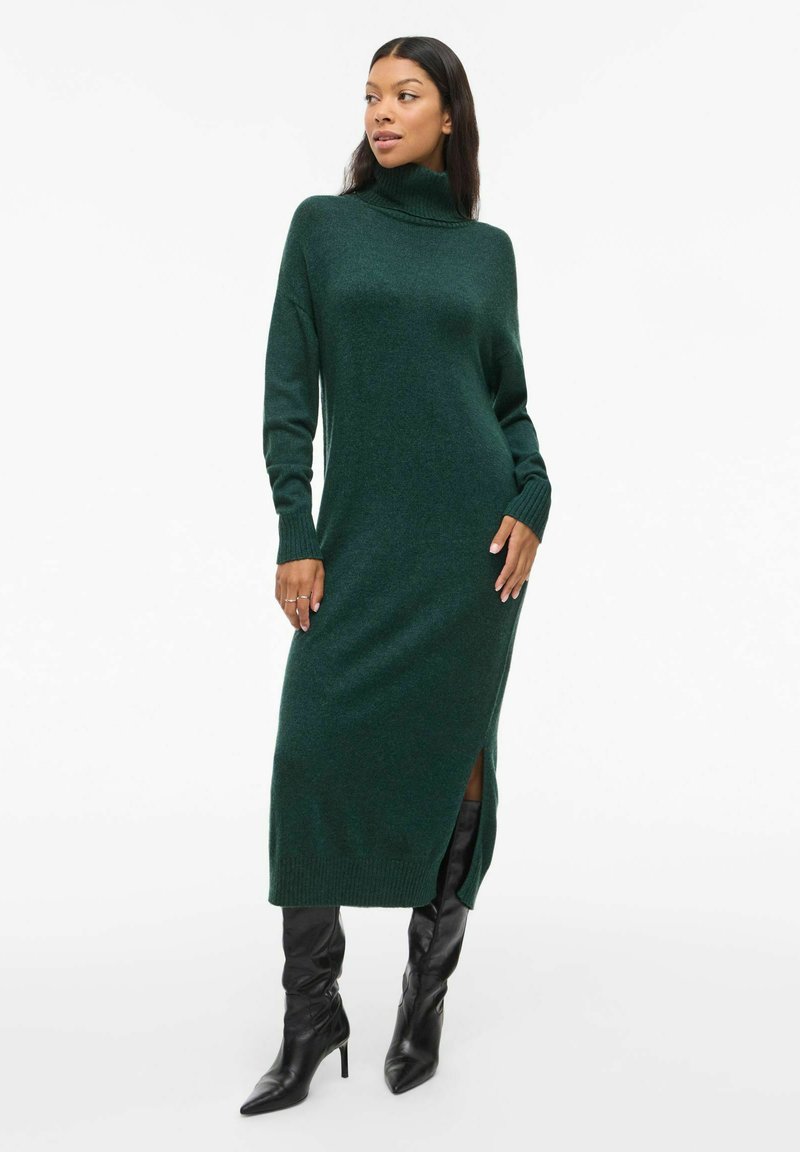 Abito lungo verde a maglia con colletto alto, polsini a costine e spacchi laterali, abbinato a stivali neri sopra il ginocchio. Tessuto liscio.