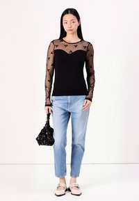 Langärmliges schwarzes Oberteil aus durchsichtiger bestickter Spitze, tailliert, kombiniert mit hellblauen Jeans und einer schwarzen Tasche. Klassische Schuhe runden den Look ab.