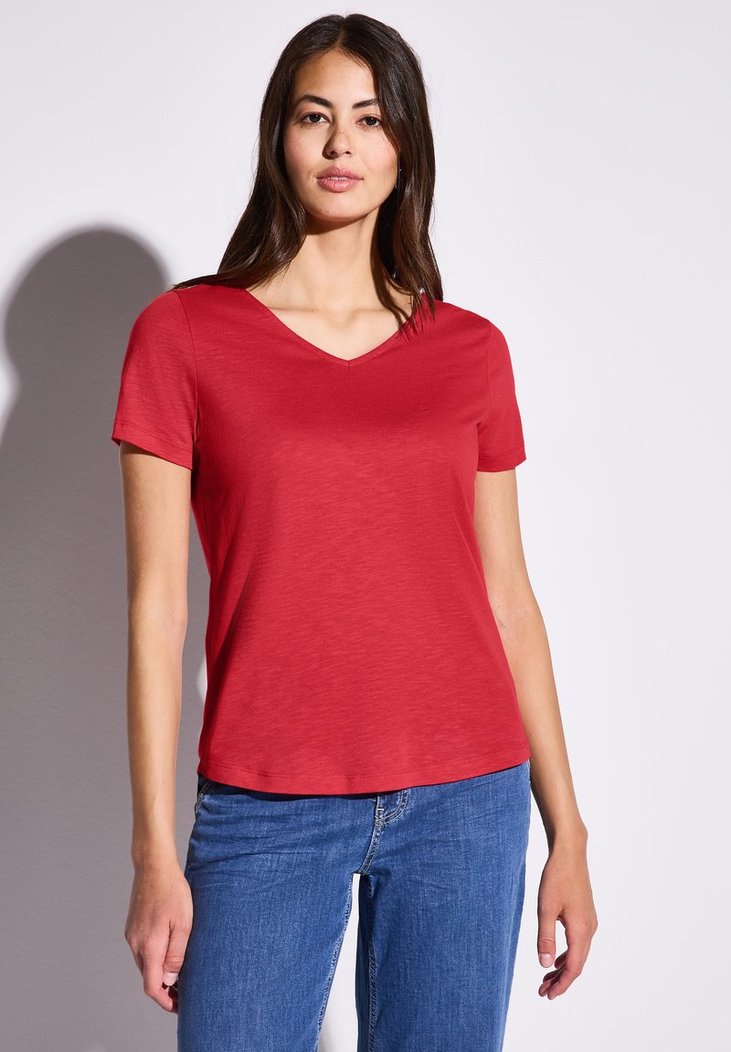 Röd V-ringad t-shirt med korta ärmar, tillverkad av ett mjukt material. Matchad med blå jeans står modellen framför en enfärgad bakgrund.