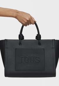 Czarna skórzana torba typu tote charakteryzuje się teksturowaną powierzchnią, wyraźnym logo "TOUS" oraz węzłowymi akcentami uchwytów. Solidny prostokątny kształt.