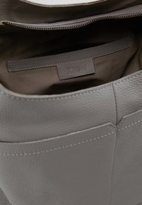 Harmaa nahkakäsilaukku, jossa on vetoketjukiinnitys, logo-tila ja tasku. Pehmeä pinta, johon on lisätty hienovaraisia ompeluyksityiskohtia.