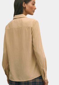 Camicia a maniche lunghe color marrone chiaro con colletto, texture liscia, polsini con bottoni e design del retro a yoke. Abbinata a pantaloni a quadri.