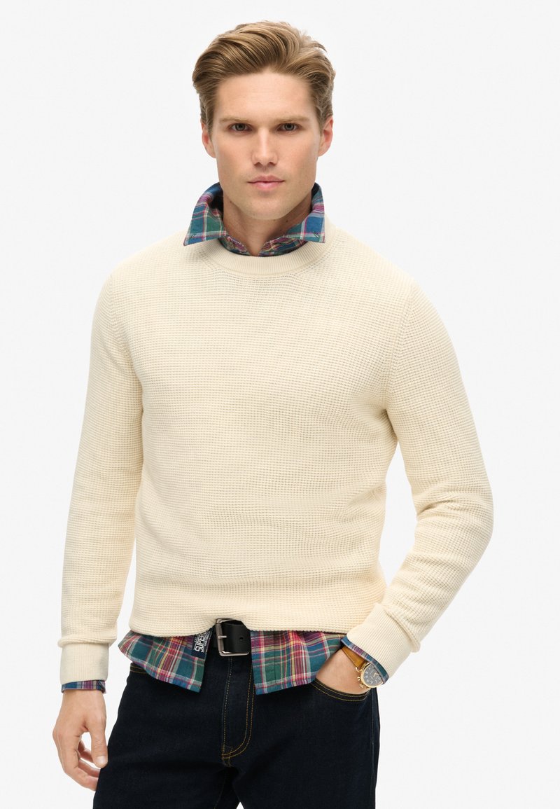 Crème textuurtrui met een ronde hals, gelaagd over een blauw-groene geruite blouse. Donkere jeans en een zwarte riem maken de outfit compleet.