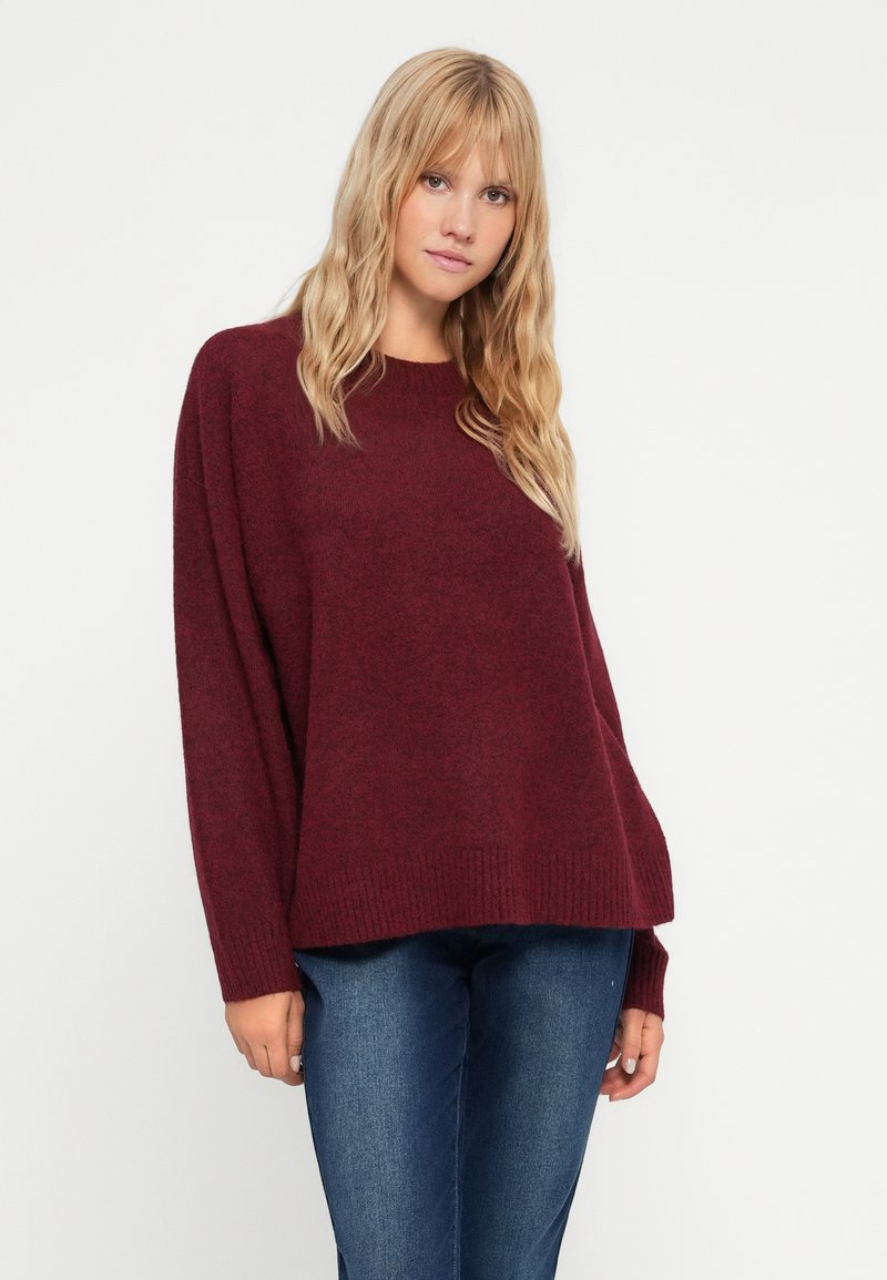 Suéter burdeos de gran tamaño con mangas largas, dobladillo acanalado y textura suave, combinado con jeans de mezclilla ajustados.