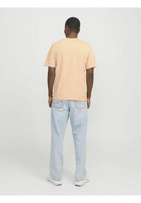Camiseta corta de manga corta en durazno claro con cuello redondo, combinada con pantalones vaqueros sueltos de lavado claro y zapatillas blancas, vista desde atrás.