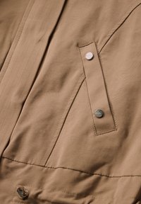 Beige takki kangas, jossa on sileä pinta. Sisältää näkyvän napsulukon ja sivutaskun, jossa on eri värinen tikkaus.