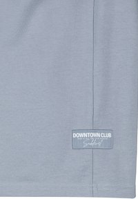 Helle blaue Stoff mit einem kleinen rechteckigen Aufnäher mit der Aufschrift "DOWNTOWN CLUB RISK IT ALL FOR YOUR PASSION Sublevel" nahe dem unteren Saum.
