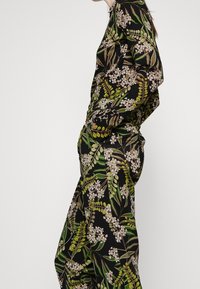 Vêtement à imprimé floral noir, avec des feuilles vertes et des fleurs blanches. Le tissu a une texture lisse, avec des manches longues et une coupe décontractée.