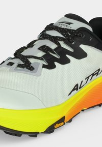 Gros plan d'une chaussure de course blanche et noire avec des lacets noirs, une semelle jaune et orange, et les logos "ALTRA" et "Vibram" visibles.