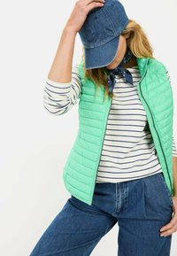 Minzgrüne, gesteppten Weste über einem weißen und navyblauen gestreiften Langarmhemd. Kombiniert mit blauen Jeans und einer Denim-Mütze. Navyfarbener Schal als Akzent.