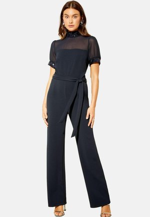 Marineblå jumpsuit med en gennemsigtig, pyntet kortærmet overdel og vide bukser, der har en bindebånd i taljen og en glat stofstruktur.