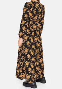 Kvinna klädd i en lång svart klänning med ett heltäckande gult blommönster och svarta kraftiga stövlar, sedd bakifrån.