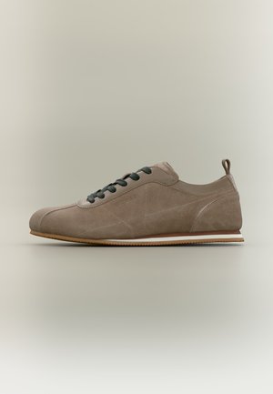 Lichtbruine suède sneaker met zwarte veters, afgeronde neus, trekkoordje bij de hiel en een platte rubberen zool met witte en bruine strepen.