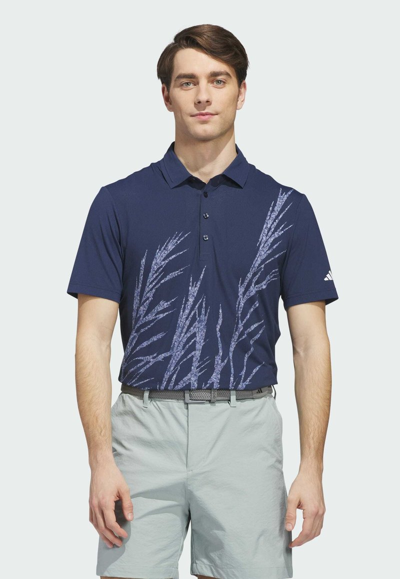 Homme portant un polo bleu marine avec un motif d'herbe bleu clair, un short gris clair et une ceinture grise, debout devant un fond blanc.