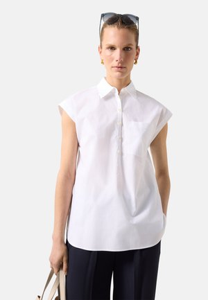 Kobieta w białej, bezrękawnikowej koszuli na guziki, czarnych spodniach, złotych kolczykach kołach, okularach przeciwsłonecznych na głowie, trzymająca beżową torebkę na jednolitym tle.