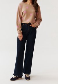 Femme portant un pull beige avec des cœurs roses, un jean large taille haute bleu marine foncé et des baskets bleu marine et blanches, posant avec une main dans la poche.