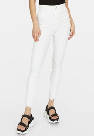 Kobieta ubrana w białe, obcisłe jeansy z wysokim stanem i czarne sandały na platformie z grubymi białymi podeszwami, stojąca na tle jednolitego tła.