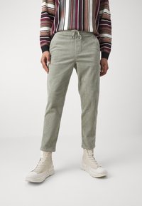 Light green corduroy pants with a drawstring waist, tapered legs, and horizontal ribbed texture, paired with light-colored boots and a striped sweater.