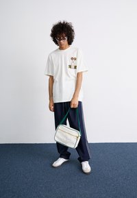 Hvid bomulds T-shirt med kaktus grafik, marineblå sweatpants med striber, hvid taske med grønne detaljer og hvide sneakers med grå såler.