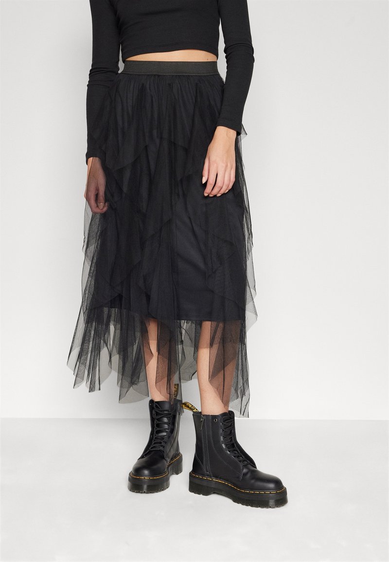 Femme portant une jupe midi en tulle noire superposée, un crop top noir et des bottines noires épaisses à lacets.