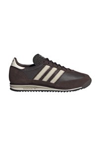 Tamno smeđe Adidas tenisice s bež prugama, suHOLJENIM naglascima, teksturiranjem jezika i čvrstim potplatima, prikazane sa desne strane na bijeloj pozadini.