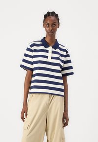 Polo a righe blu e bianche con colletto, maniche corte e tre bottoni. Indossato con pantaloni cargo beige.