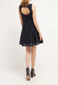 Robe sans manches bleu marine en tissu texturé, corsage ajusté, jupe évasée et détail de dos en forme de trou de serrure. Sandales à talons hauts noires.