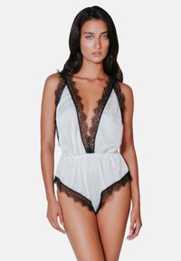 Vit bodysuit med spetskant, djup V-ringning, justerbara axelband och samlad midja med svarta spetsdetaljer längs kanterna.