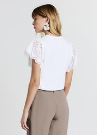 T-shirt blanc en coton avec manches en dentelle ajourée brodée. Les manches présentent des bords festonnés. Associé à un pantalon beige taille haute.