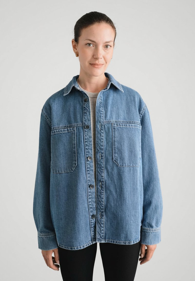 Kobieta z ciemnymi włosami w koku, ubrana w luźną niebieską dżinsową kurtkę na szarym podkoszulku i czarnych spodniach, stojąca na tle jasnego, jednolitego tła.