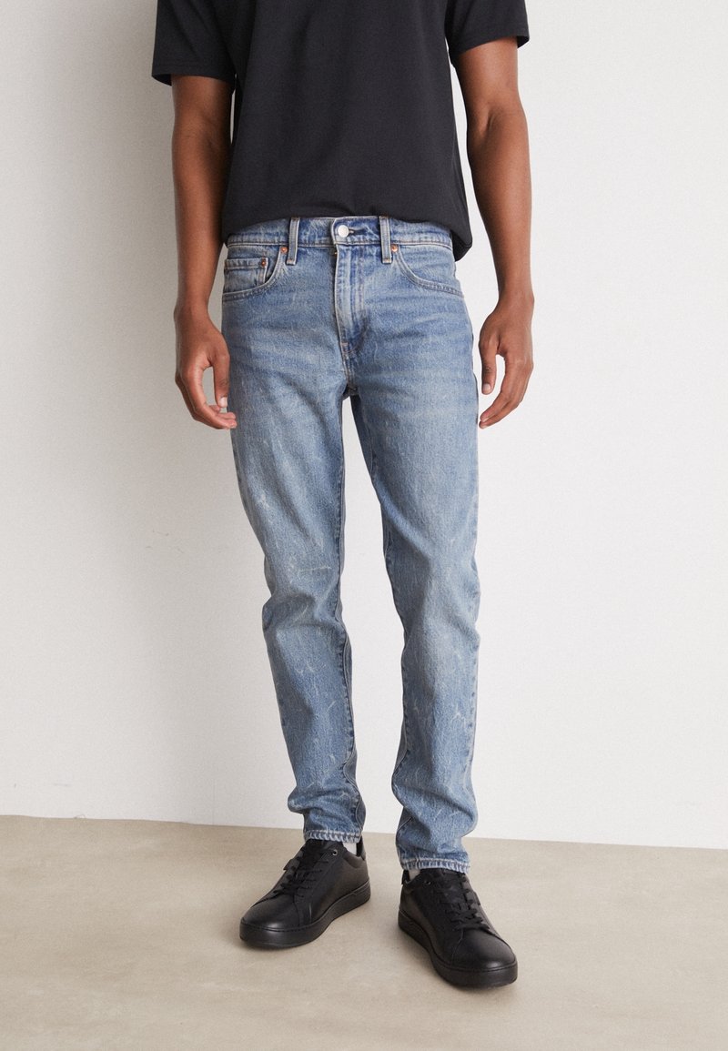 Personne portant un jean slim bleu clair, un t-shirt noir et des baskets noires, debout sur un sol gris contre un mur blanc.