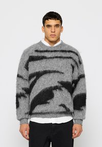 Γκρι πουλόβερ από mohair με μαύρα αφηρημένα σχέδια, στρογγυλή λαιμόκοψη, μακριά μανίκια και ριμπ μανσέτες, φορεμένο πάνω από ένα λευκό πουκάμισο.