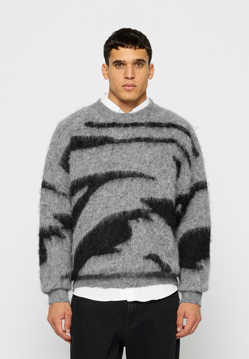 Γκρι πουλόβερ από mohair με μαύρα αφηρημένα σχέδια, στρογγυλή λαιμόκοψη, μακριά μανίκια και ριμπ μανσέτες, φορεμένο πάνω από ένα λευκό πουκάμισο.