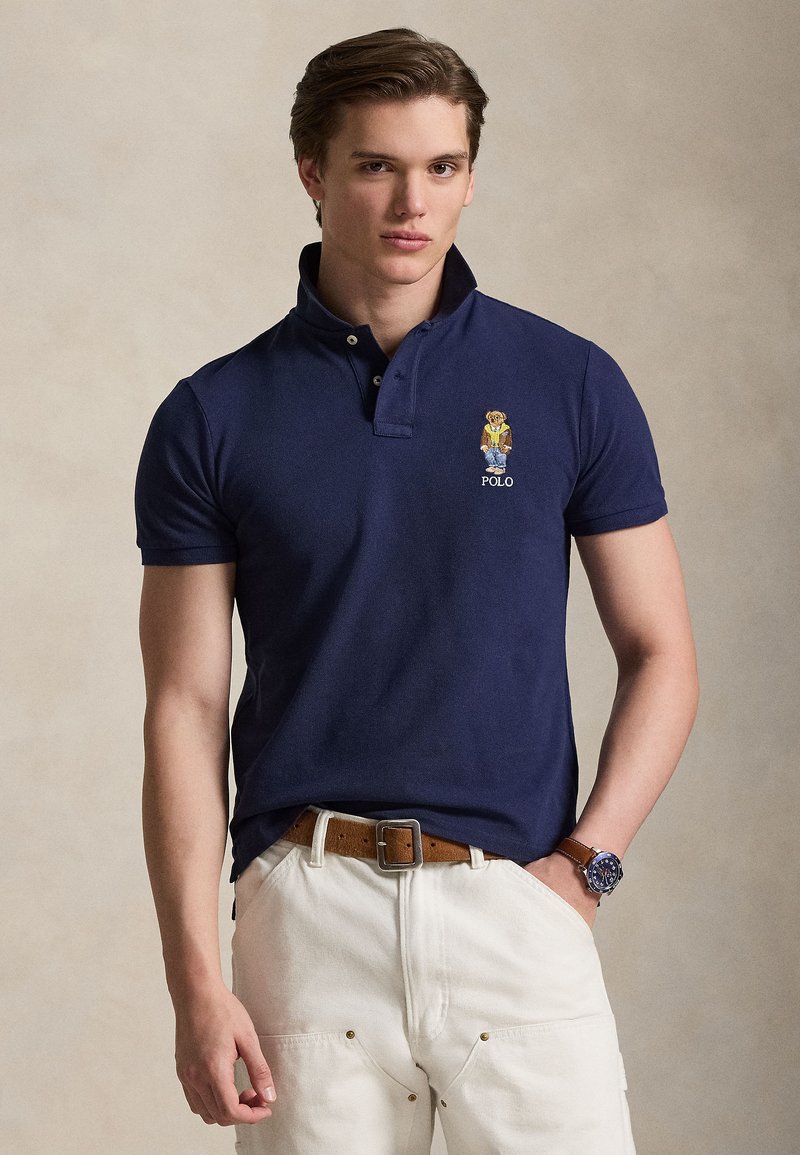 Polo bleu marine en tissu texturé avec une patte de boutonnage à deux boutons, logo ours brodé et coupe décontractée, porté avec un pantalon blanc.