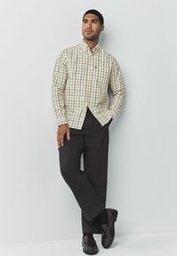 Camisa a cuadros beige con cuello abotonado, mangas largas y un bolsillo en el pecho, combinada con pantalones negros y mocasines oscuros.