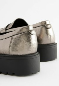 Zapatos deslizables metálico plata con parte superior texturizada, acentos de costura a juego y suelas de goma negra gruesa. Parte trasera redondeada y detalles de diseño mínimos.