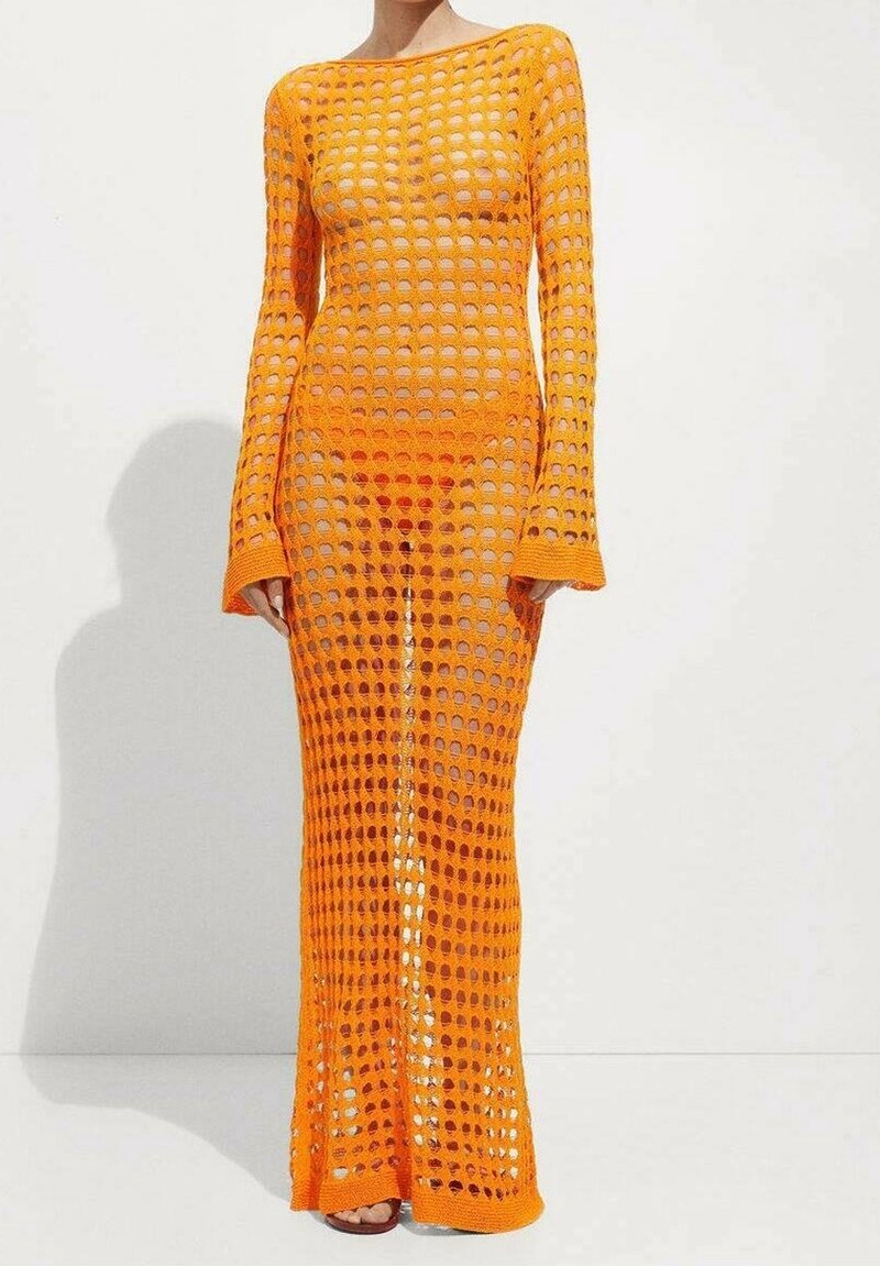 Femme portant une longue robe au crochet orange vif avec de grands trous et des manches longues, se tenant devant un fond blanc uni.