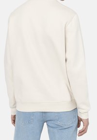 Felpa beige chiaro con colletto rotondo, polsini e orlo a coste; tessuto morbido con una texture liscia; indossata con jeans in denim blu chiaro.