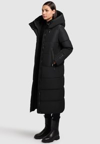 Caban lung puffer negru cu glugă, cu închidere prin fermoar, buzunare laterale, design matlasat și o textură netedă. Purtați-l cu cizme negre.
