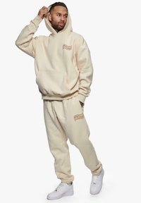 Beige hettegenser med frontlomme og hette, kombinert med matchende joggebukser. Har oransje logo på buksene og hvite joggesko. Mykt materiale.