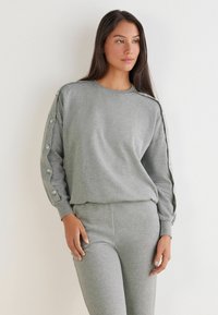 Grå sweatshirt med lange ærmer, der har dekorative knapper på skuldrene, parret med matchende grå bukser. Blødt stof og afslappet pasform.