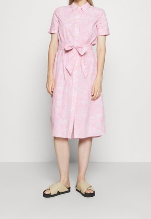 Kvinde iført en pink mønstret midi skjortekjole med korte ærmer og bånd i taljen, kombineret med beige flade sandaler med krydsremme.