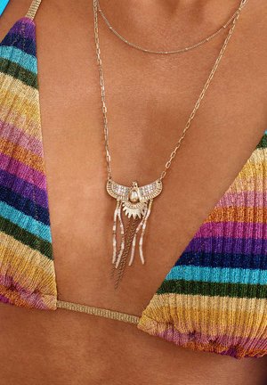 Collier en ton doré avec un pendentif en forme d'oiseau orné de pierres transparentes et de perles blanches, complété par des chaînes fines et un tissu rayé coloré.